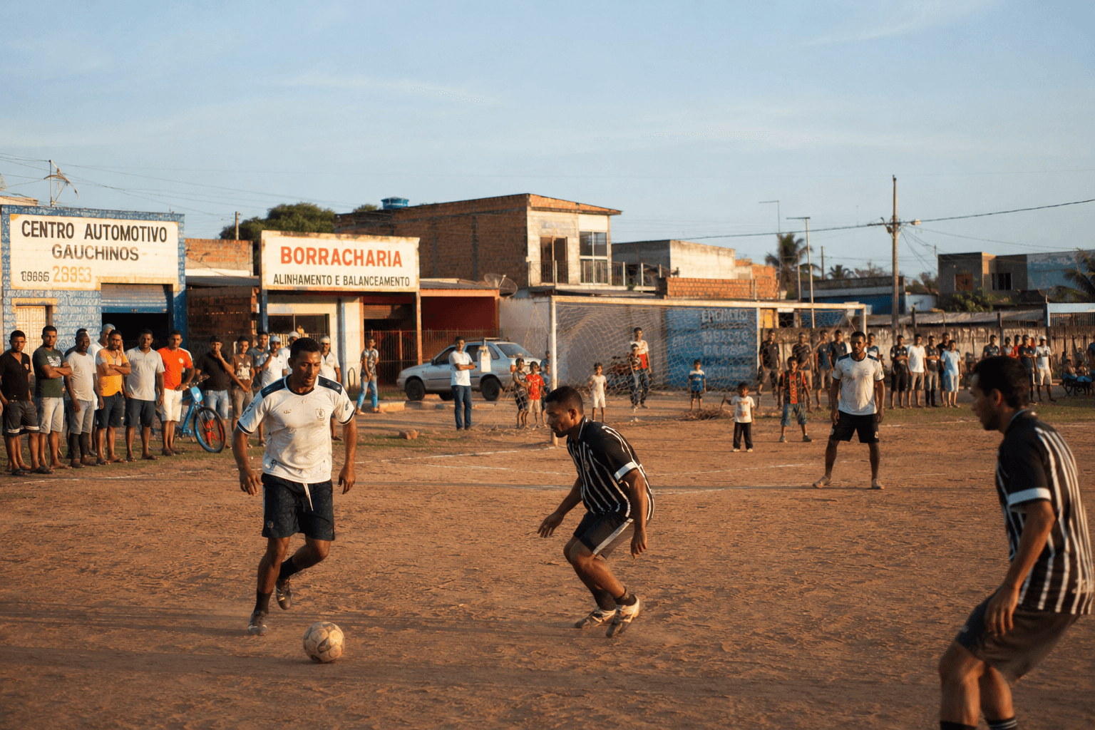 Time de futebol de várzea em campo, com torcida e comércios locais ao fundo